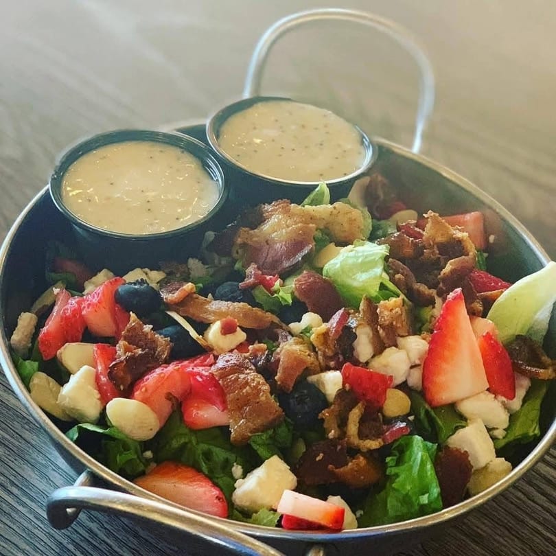 A vibrant salad with strawberries, blueberries, bacon, and feta cheese, served with two dressings.
