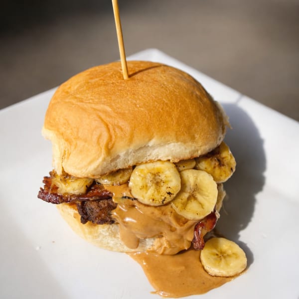A peanut butter, bacon, and banana sandwich on a bun, with peanut butter dripping onto a white plate.