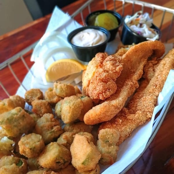 Fried fish and okra with coleslaw, tartar sauce, and pickles in a metal basket.