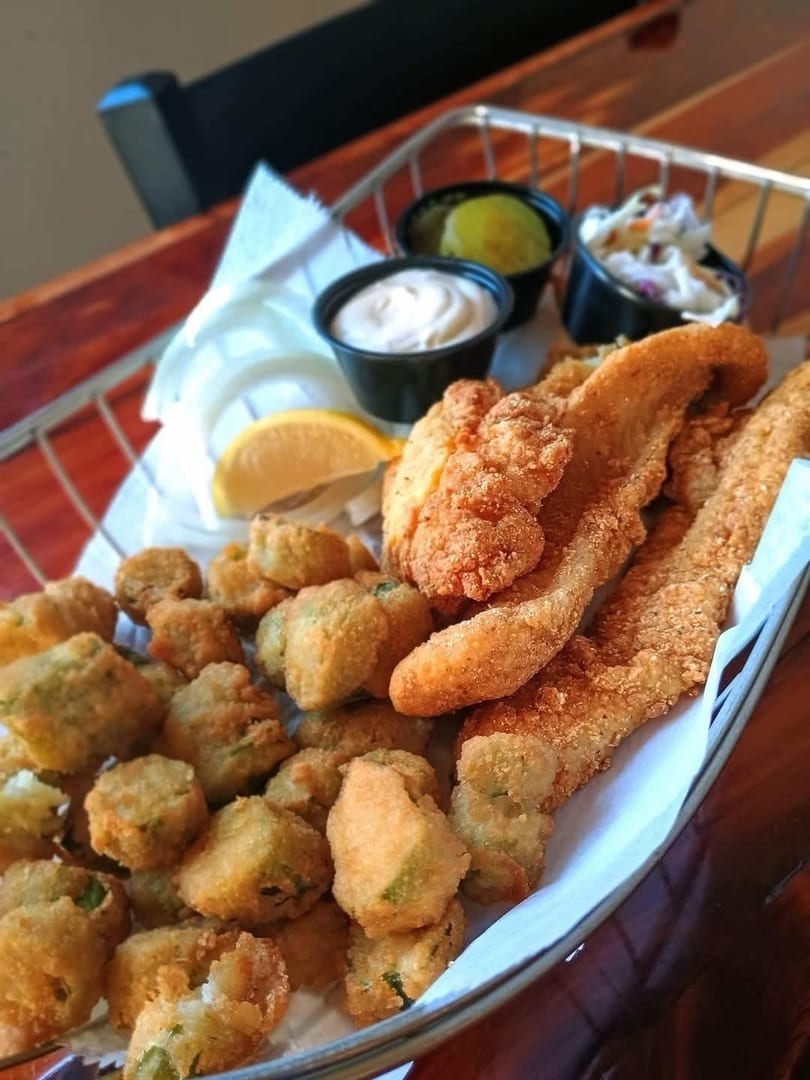 Fried fish and okra with coleslaw, tartar sauce, and pickles in a metal basket.